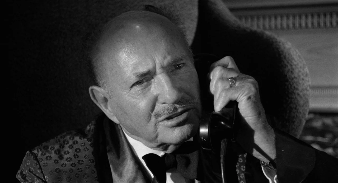 A Face In The Crowd (1957) movie still: closeup — An elderly man in formal attire is situated in a dark room. He is speaking into a black vintage…