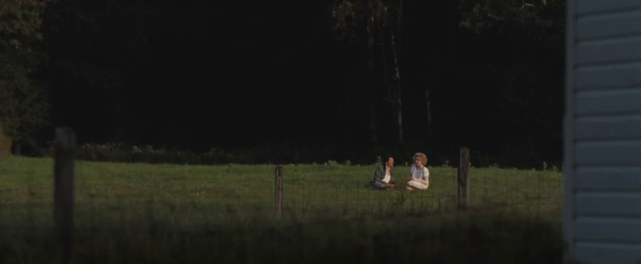 Martha Marcy May Marlene (2011) movie still: wide — An open grassy field sits bordered by a dark dense forest line. A man and a woman sit together…