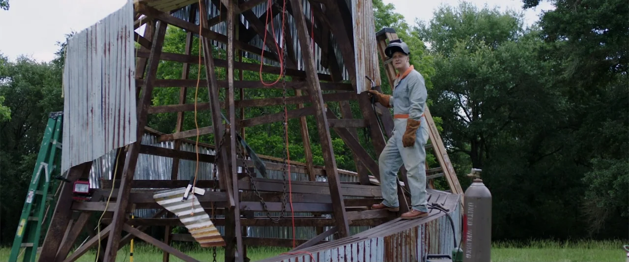 Playing God (2021) movie still: fullbody — A wooden scaffold structure under construction stands in a grassy field with trees behind. A man wearing…