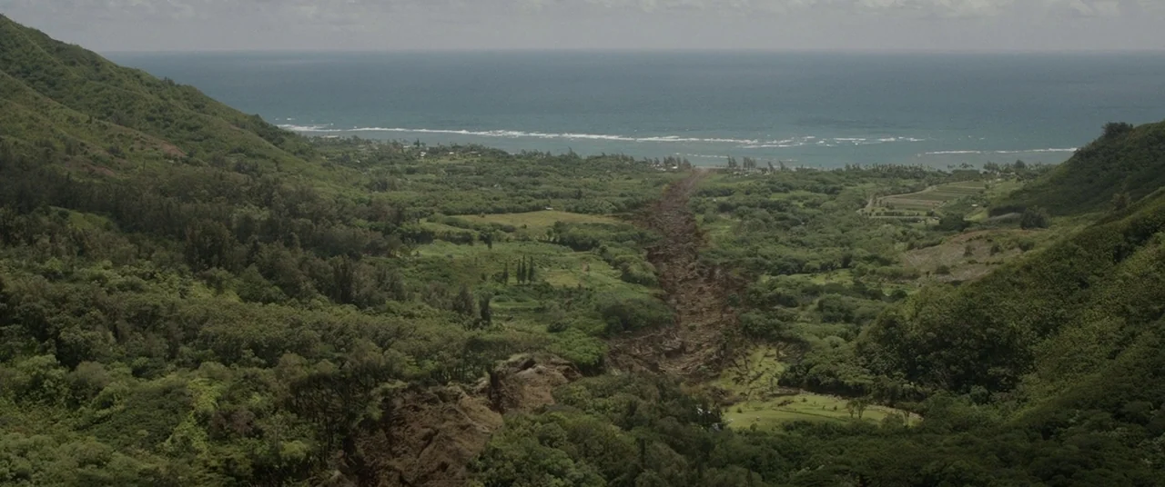 Godzilla (2014) movie still: establishing — A lush tropical valley stretches toward a coastline with a long, dark earthen fissure. The dense green…