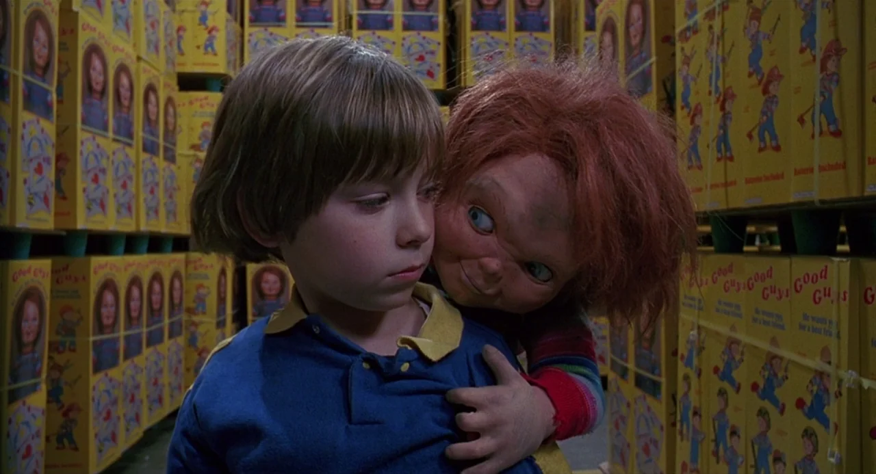 Child’s Play 2 (1990) movie still: medium — A young child stands surrounded by stacks of boxed dolls in a dim warehouse. The Good Guy doll leans…