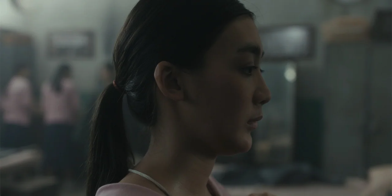 Host (2020) movie still: closeup — A dimly lit interior space features metal lockers and blurred figures in the background. A young woman with her…