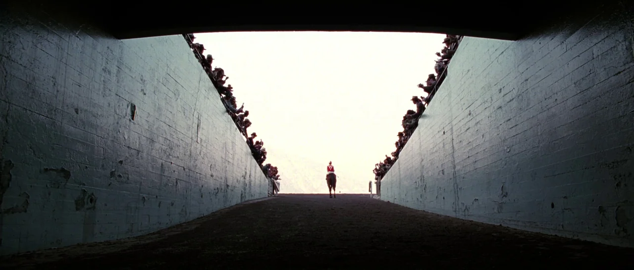 Seabiscuit (2003) movie still: wide — A long concrete tunnel provides a view toward a bright outdoor horizon. A lone jockey on a horse walks away…