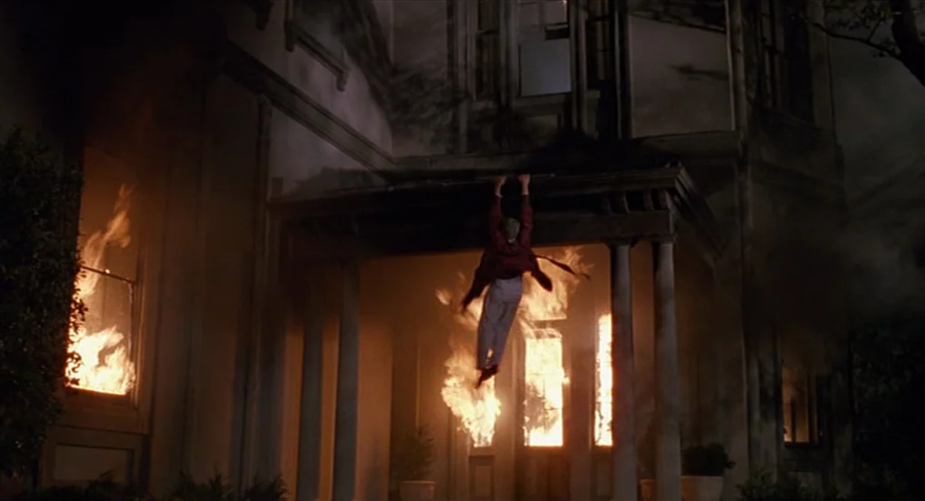 Miller’s Crossing (1990) movie still: wide — A classic two-story house at night with flames erupting from the windows. A person hangs suspended from…