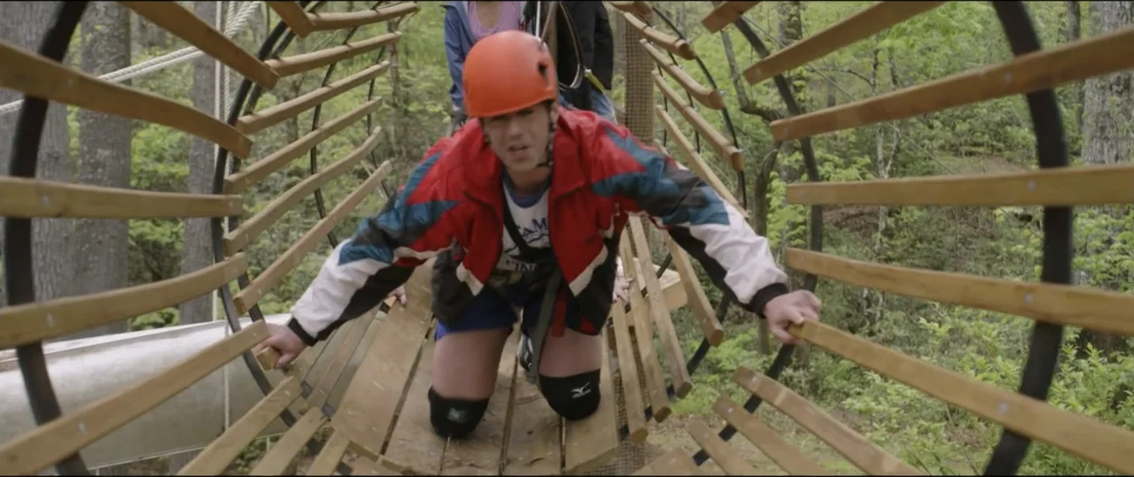 Summer Camp (2024) movie still: medium — A wooden barrel-shaped bridge is suspended high in a lush green woodland area. A man in a climbing helmet…