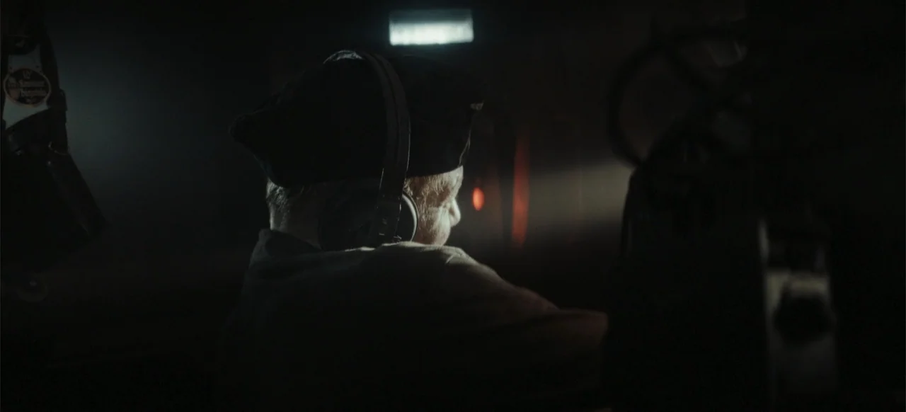 The Tank (2025) movie still: medium — A dimly lit, cramped cabin is filled with shadows and industrial metal surfaces. A man wearing headphones…