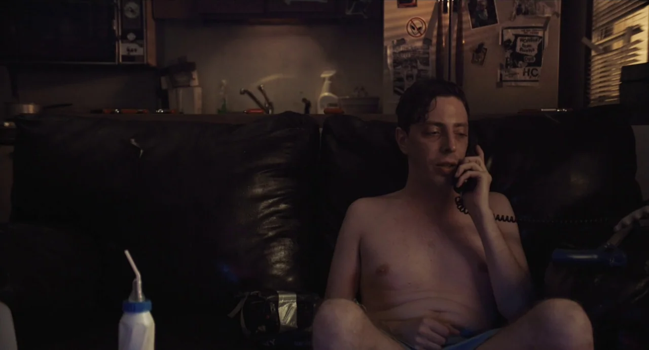 Relaxer (2018) movie still: medium — A dark living room contains a black leather couch in the foreground. A shirtless man sits on the couch while…
