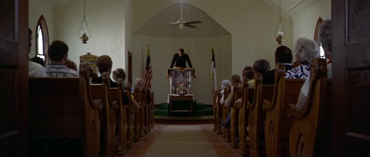Thunderbolt & Lightfoot (1974) movie still: wide — A modest church interior features rows of dark wooden pews facing a central pulpit. A man in a…