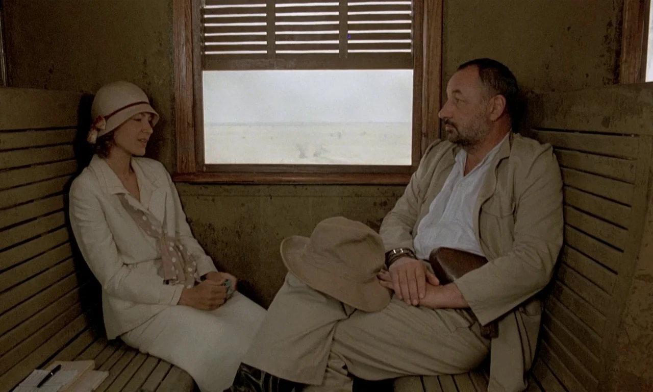 Coup De Torchon (1981) movie still: medium — A rustic train compartment features wood-slatted walls and a view of a desert landscape. A man and…