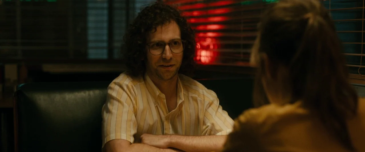 Brigsby Bear (2017) movie still: medium — A dark booth in a vintage diner is illuminated by a nearby neon sign. A man with curly hair and glasses…