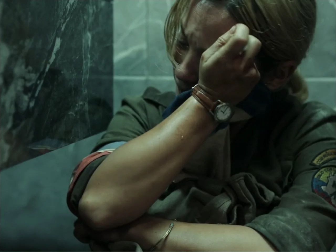 November (2025) movie still: closeup — A dark, cramped space with tiled walls and poor lighting conditions. A woman in a military uniform leans…