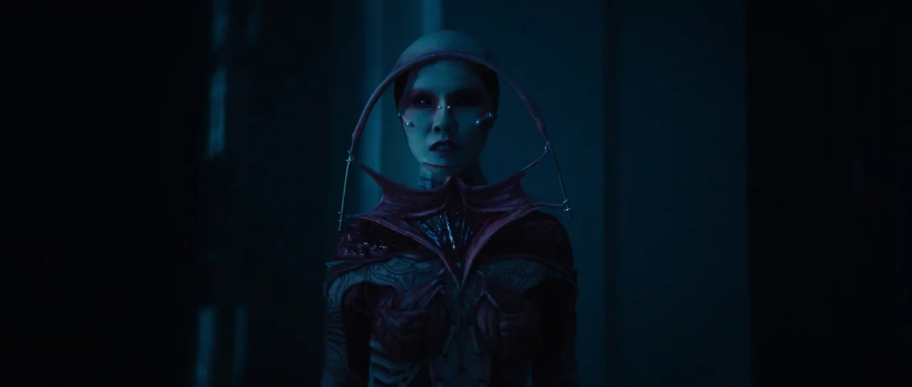 Hellraiser (2022) movie still: medium — A dark space features a figure with an organic, flesh-textured, bio-mechanical collar frame. The subject…