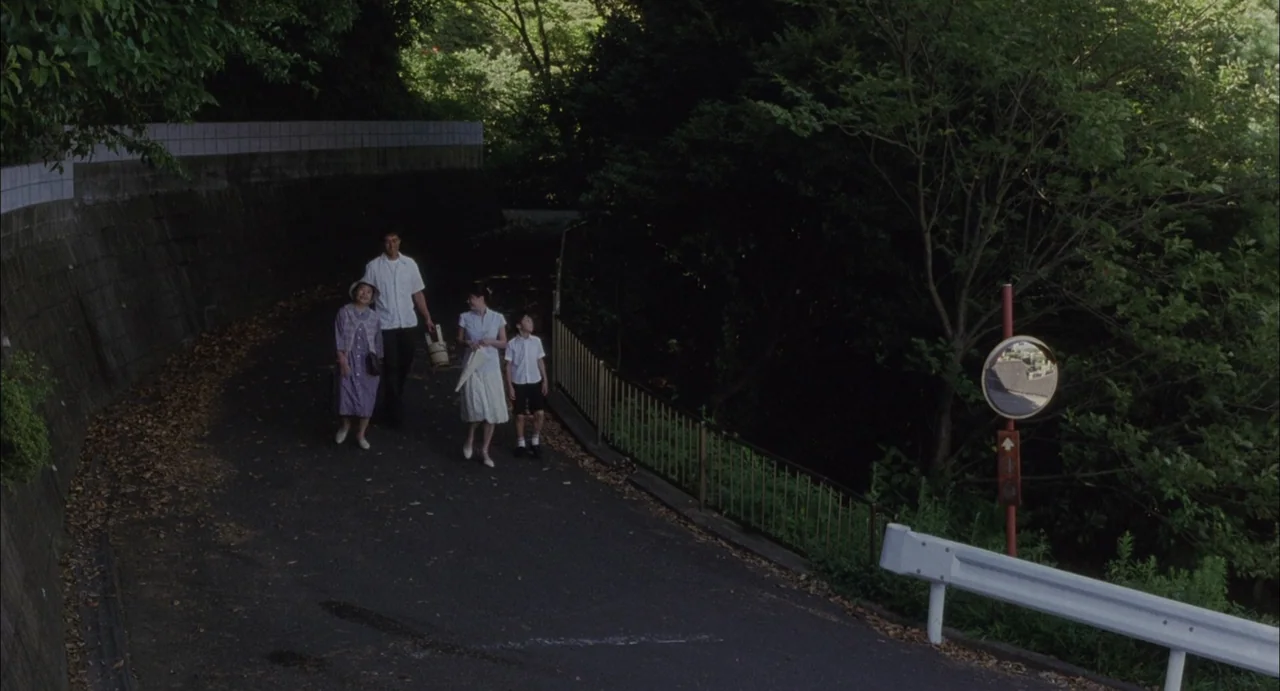 Still Walking (2008) movie still: wide — A group of four people walk down a paved path flanked by trees and greenery. A traffic safety mirror is…