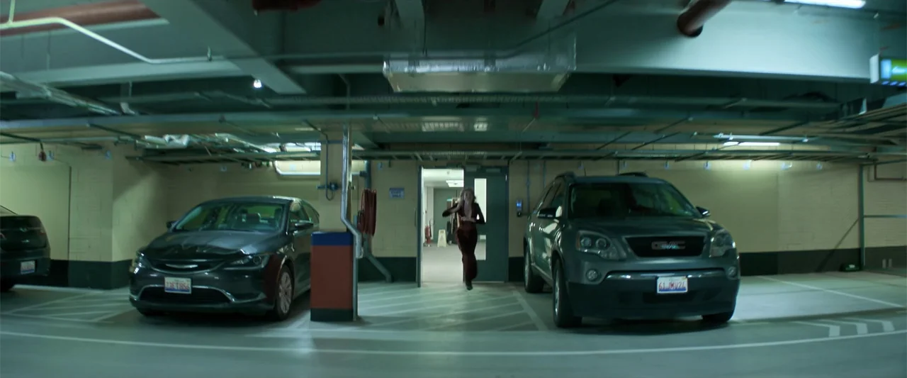 Drop (2025) movie still: wide — A utilitarian concrete parking garage features two parked vehicles flanking a central open doorway. A lone woman in…