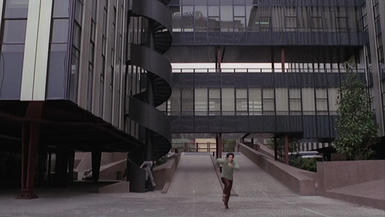 The Black Belly of the Tarantula (1971) movie still: wide — A modern brutalist building features a dark metal spiral staircase and concrete ramps. A…