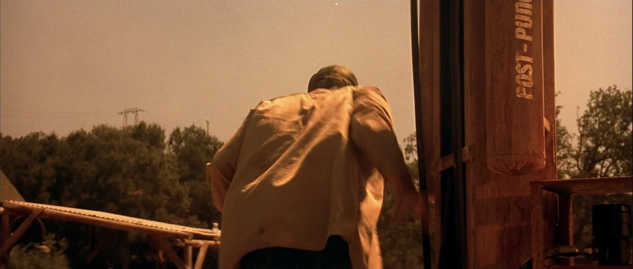 Jeepers Creepers 2 (2003) movie still: medium — An outdoor gas station lot with a rusted pump structure under a golden sky. A man seen from the back…