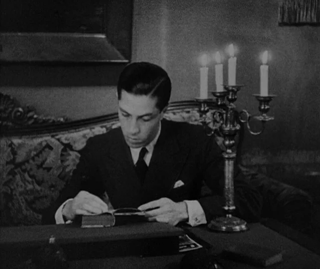 Vampyr (1932) movie still: medium — A dark study contains a man sitting at a desk with an ornate candelabra. The man writes in a small ledger while…