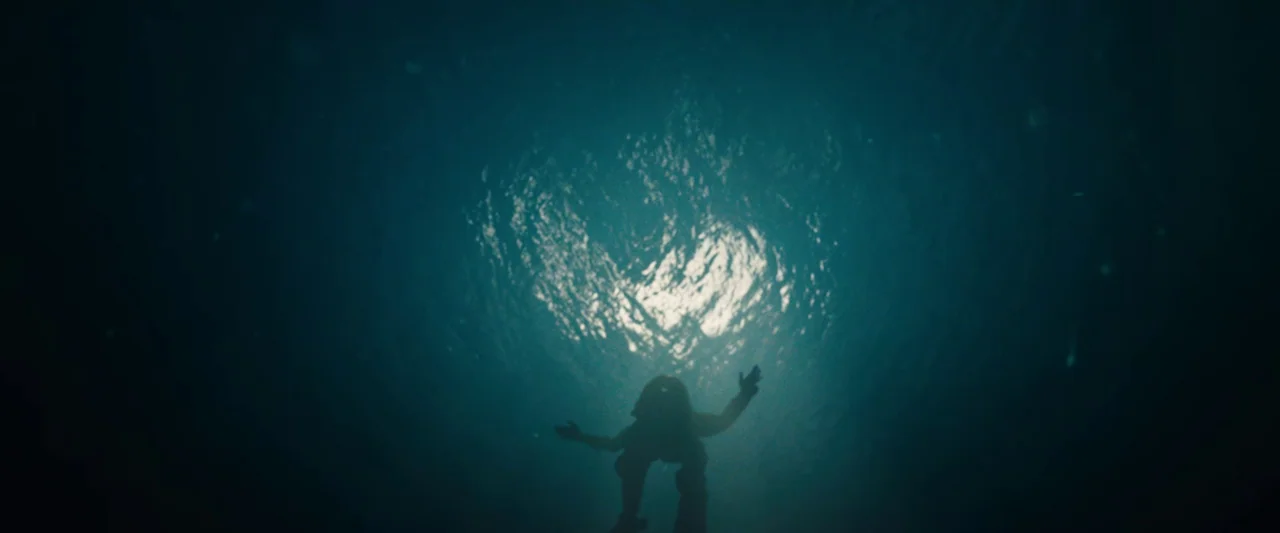 Black Sea (2014) movie still: wide — A person floats in deep, dark ocean water beneath a bright surface reflection. The subject is seen in…
