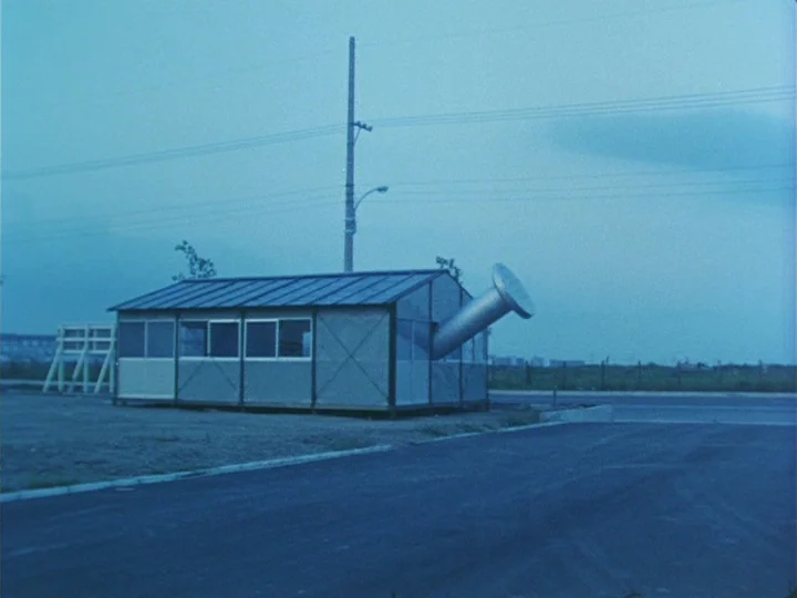 Trial (1975) movie still: wide — A modular rectangular structure with a large metal funnel sits in an open field. Utility poles and dark asphalt…