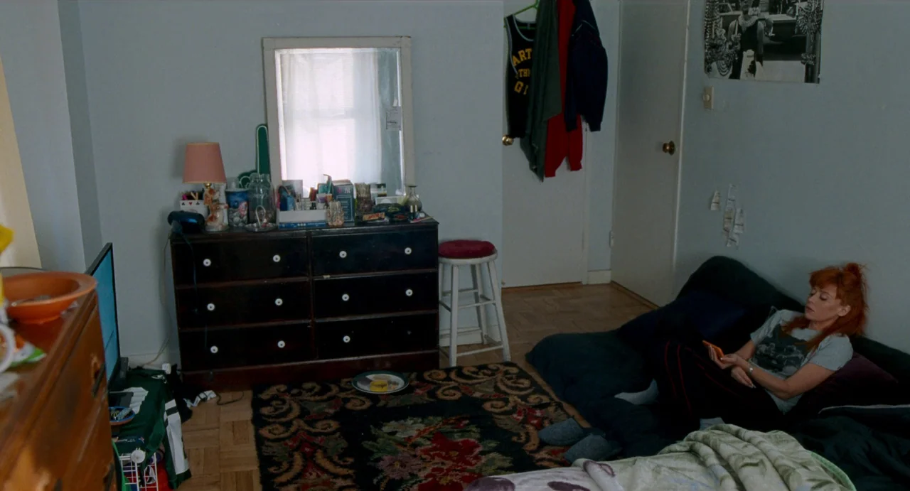 His Three Daughters (2023) movie still: wide — A cluttered bedroom has a dark wooden dresser and an ornate patterned rug. A woman rests on a bed…