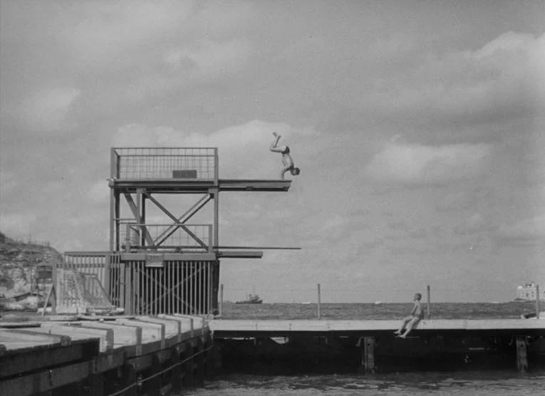 Wings (1966) movie still: wide — A wooden pier extends into the sea under a cloudy sky. A man performs a handstand on a tall metal diving structure…
