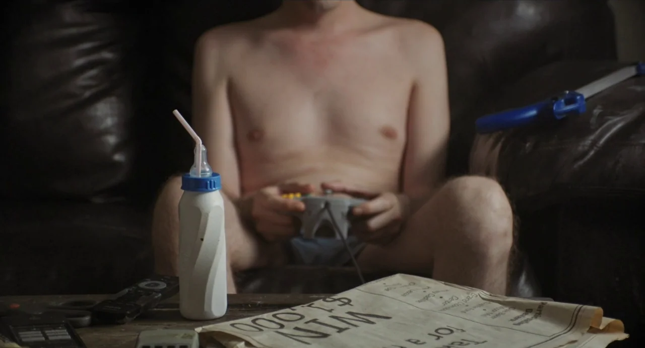Relaxer (2018) movie still: medium — A dark leather sofa sits in a room with a wooden table in front. A white baby bottle with a straw sits on the…