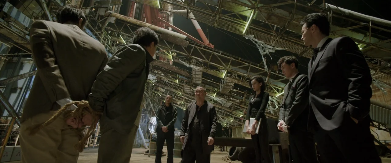 Skiptrace (2016) movie still: fullbody — A decrepit industrial hangar filled with metal scaffolding and debris under dim lighting. A group of…