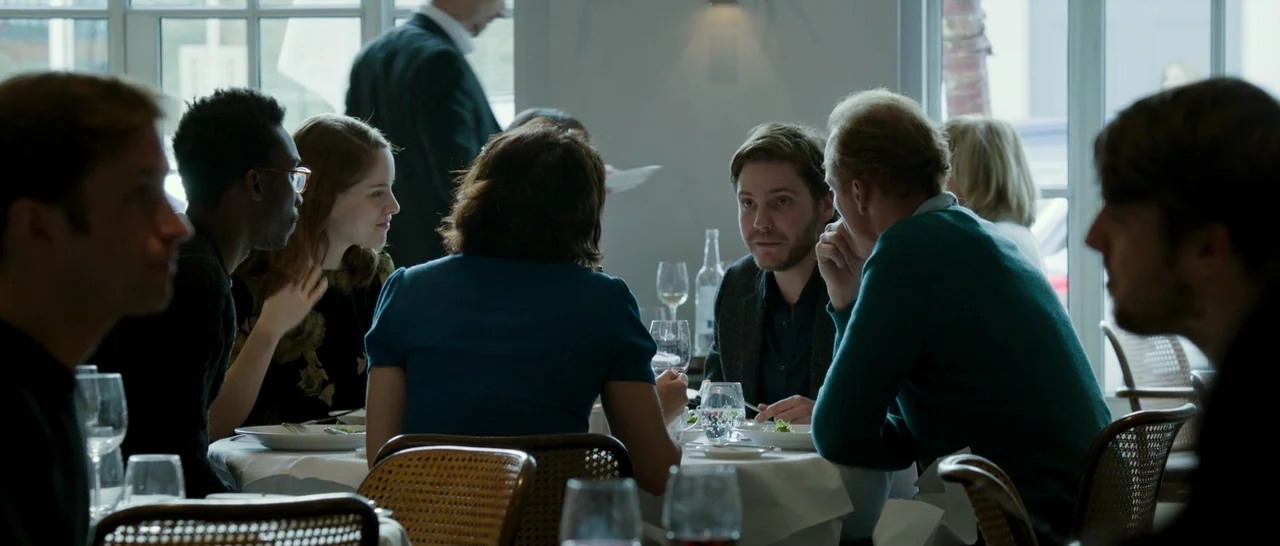 The Face of an Angel (2014) movie still: medium — A group of people sit around a table in a bright, modern restaurant setting. They are having a…