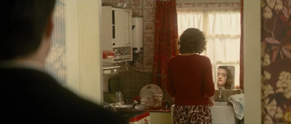 Nowhere Boy movie still: medium — A domestic space with floral wallpaper and patterned curtains near a small window. A woman stands by the basin…