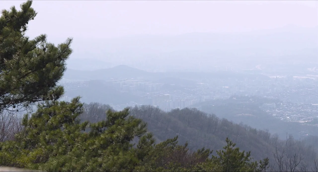 Nobody’s Daughter Haewon (2013) movie still: establishing — A hazy panoramic view looks out from a mountain top over a city. Pine branches occupy…