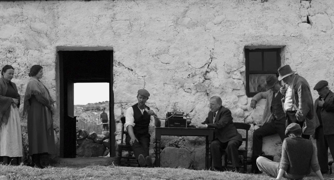 Song of Granite (2017) movie still: fullbody — A group of people gather in front of a weathered whitewashed stone cottage wall. Men and women stand…