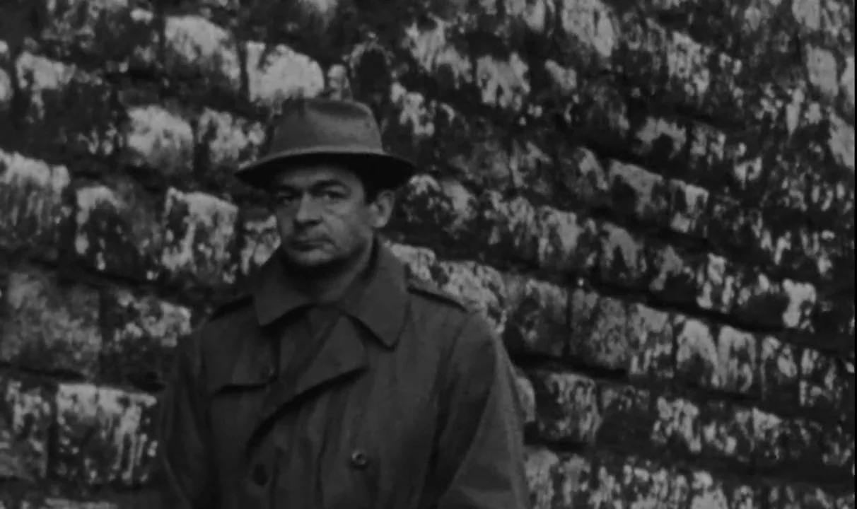 Le Doulos (1962) movie still: medium — A man stands before a weathered stone block wall with mossy grout lines. He wears a fedora and trench coat…