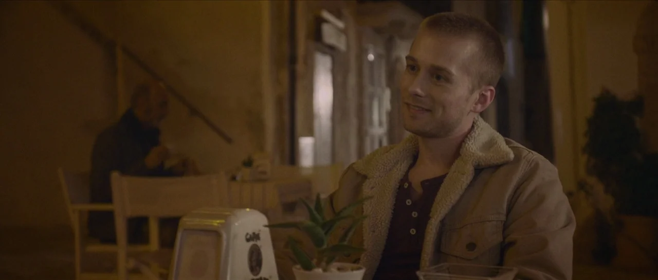 Spring (2014) movie still: medium — A dimly lit outdoor patio at night features tables arranged on a paved surface. A man with a short haircut sits…