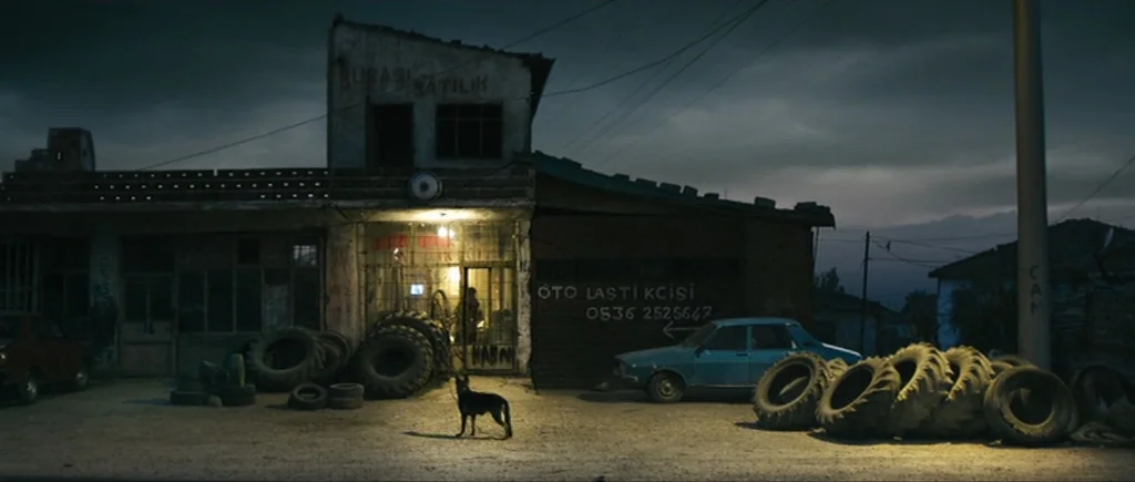 Once Upon a Time in Anatolia (2011) movie still: wide — A roadside auto repair shop at night features scattered tires and a dark facade. A lone dog…