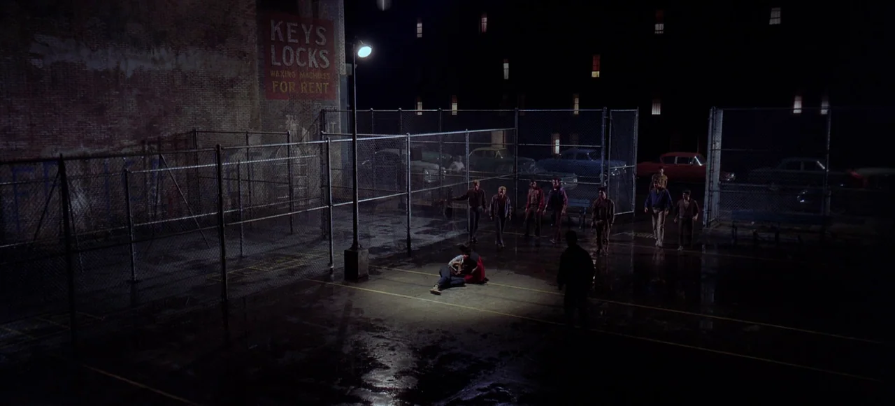 West Side Story (1961) movie still: wide — A dark, wet asphalt basketball court is enclosed by chain-link fencing at night. A group of men stands…