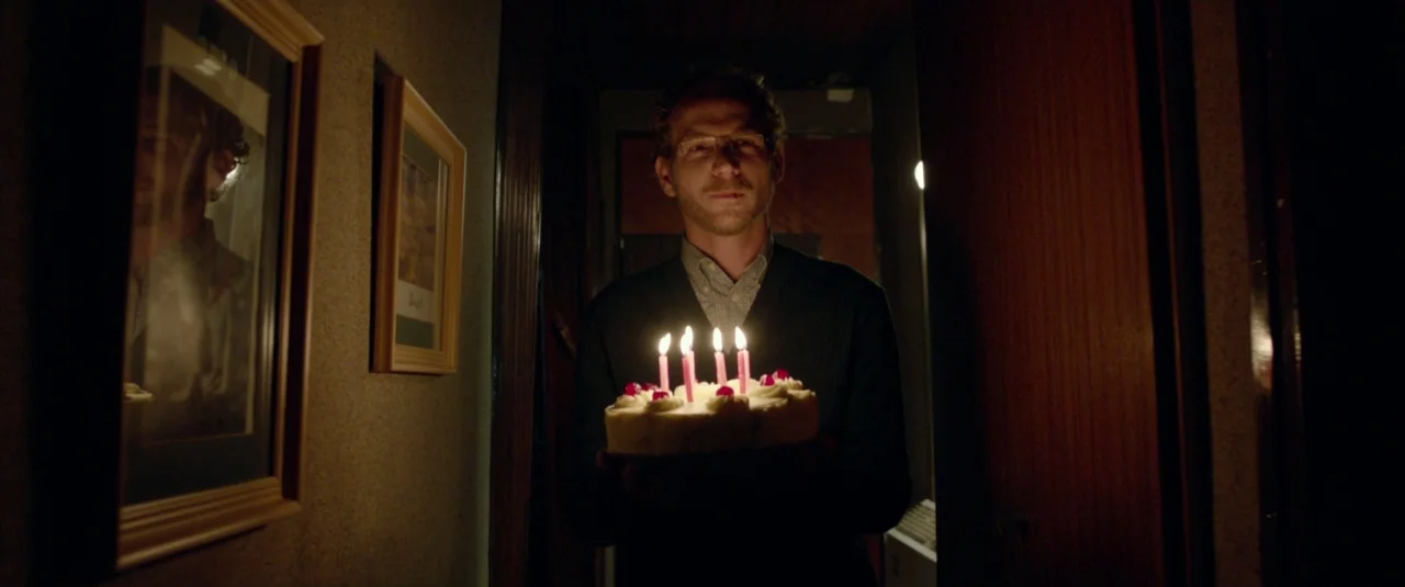 Big Bad Wolves (2013) movie still: medium — A man stands in a dark hallway holding a white birthday cake with lit candles. Framed pictures hang on…