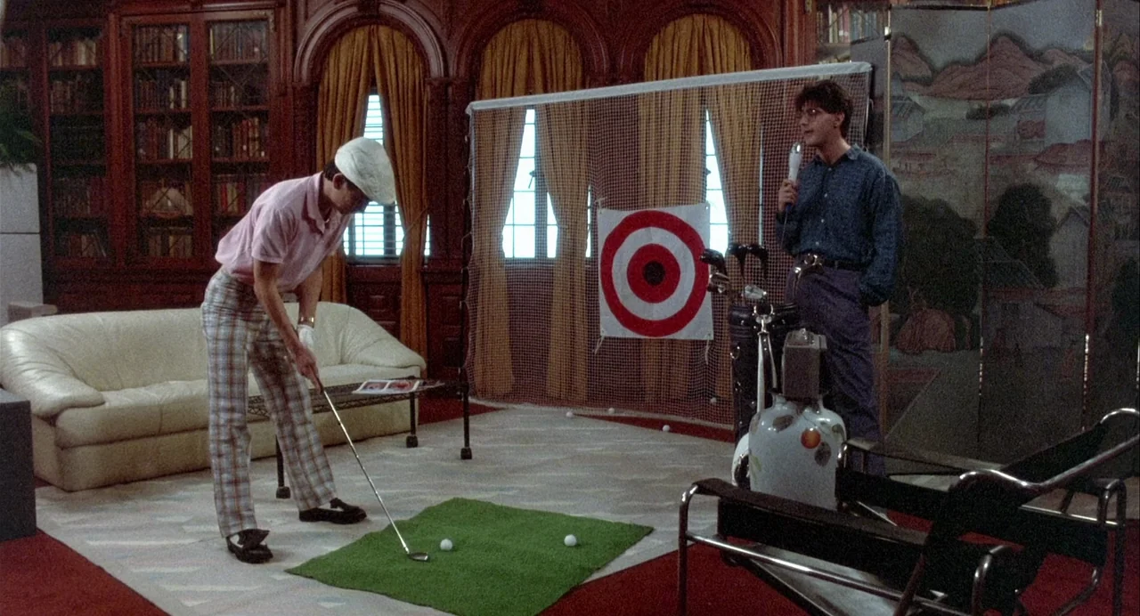 Vamp (1986) movie still: fullbody — A spacious wood-paneled room features a white leather sofa and a golf practice area. One man in a flat cap putts…