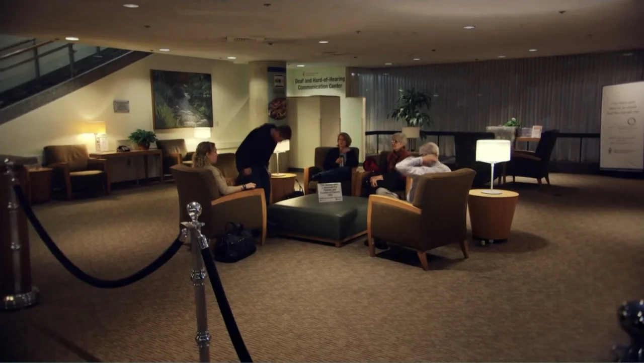 Suited (2016) movie still: wide — A spacious waiting area features several armchairs, a central ottoman, and beige carpeting. Four adults are…