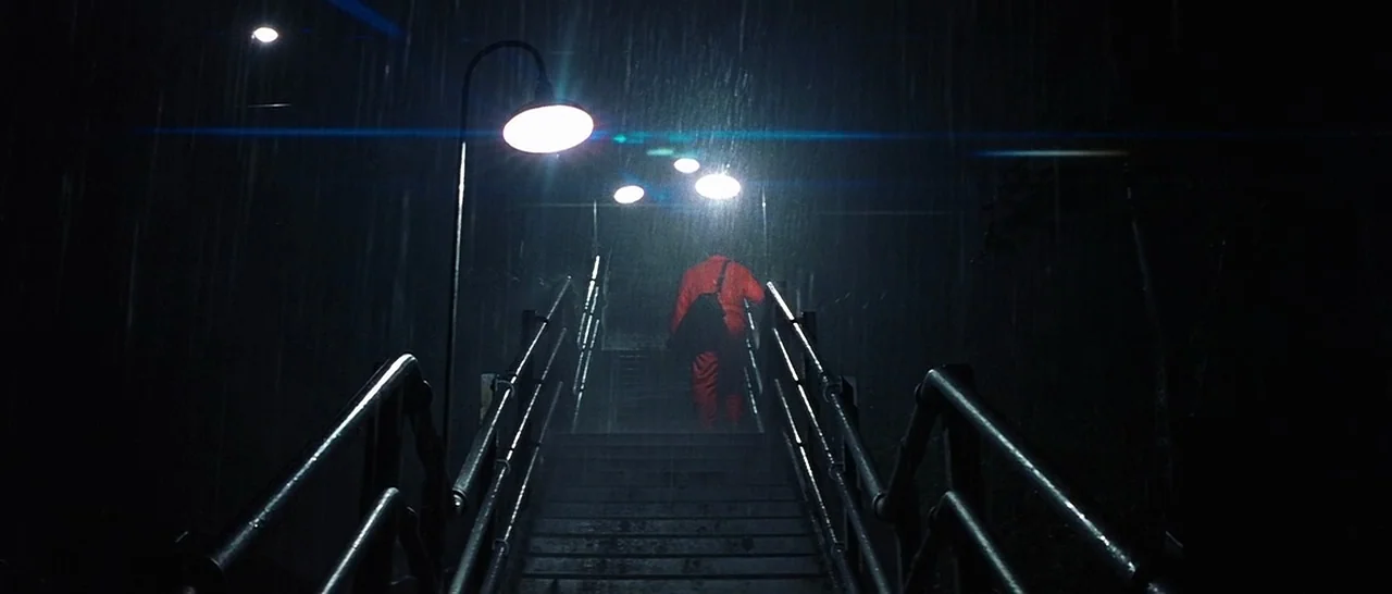 Unbreakable (2000) movie still: fullbody — A dark outdoor staircase illuminated by overhead streetlamps during a heavy rainstorm at night. A lone…