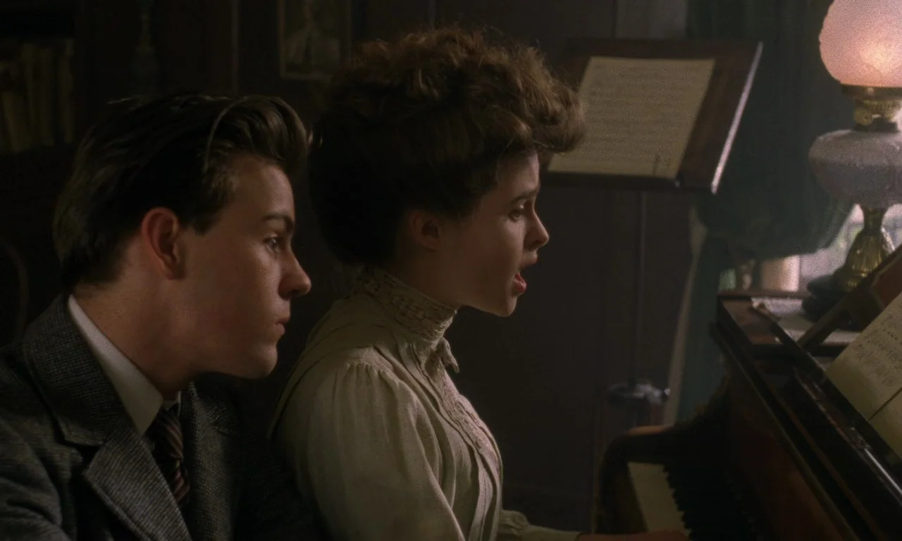 A Room With A View (1985) movie still: medium — A dimly lit room with Victorian-era decor features a piano and music stand. A young woman plays and…