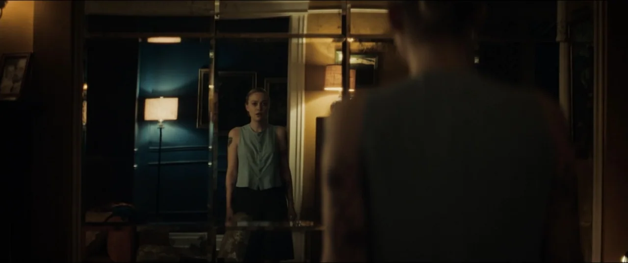 Vicious (2025) movie still: medium — A dimly lit room contains a large tiled mirror reflecting a young woman. The woman stands before the mirror…
