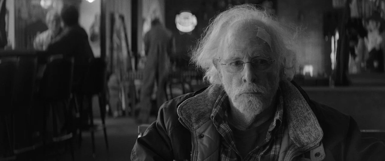 Nebraska (2013) movie still: medium — A dimly lit diner interior with bar stools and blurry figures in the background. An older man with a bandage…