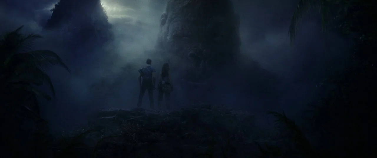 Kong: Skull Island (2017) movie still: wide — A dark, fog-shrouded tropical landscape features a sheer cliff edge. Two people stand together on the…