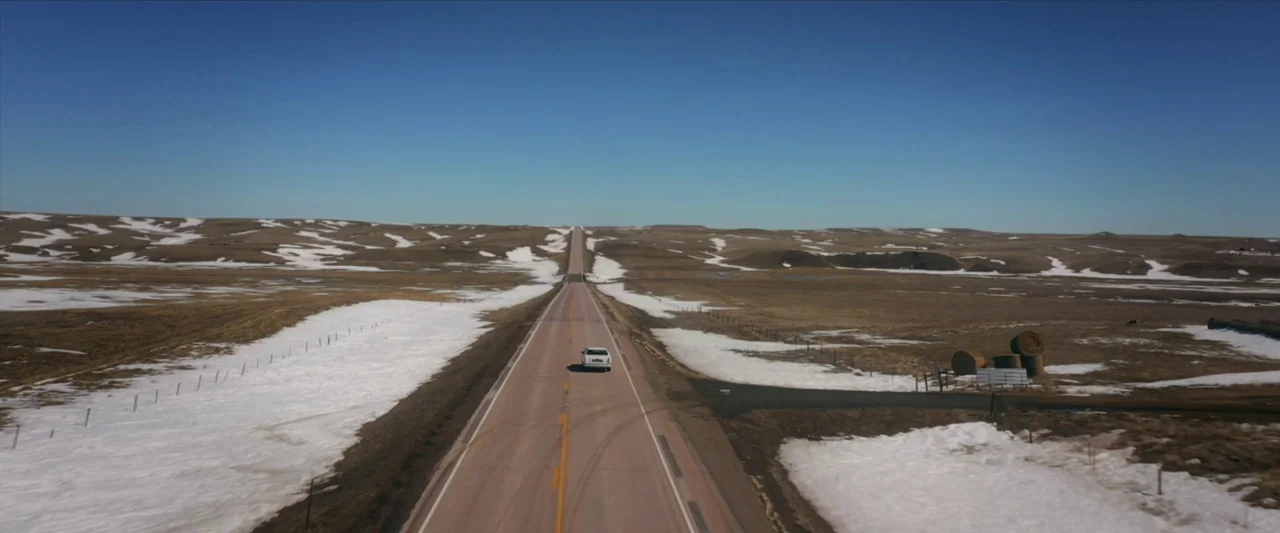 The Unknown Country (2022) movie still: wide — A vast, rolling prairie landscape features patches of remaining winter snow and dry grass. A lone…