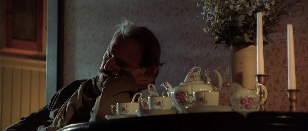 Open Range (2003) movie still: medium — A man sits at a table in a dimly lit room with floral wallpaper. He rests his face in his hand beside a…