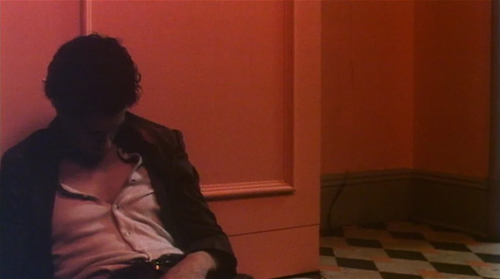Union City (1980) movie still: medium — A man slumps against an orange wall in a dimly lit hallway. The floor features a classic diamond-pattern…