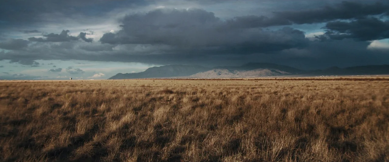 Intrusion (2021) movie still: establishing — A vast, flat expanse of dry, golden-brown grass stretches toward a distant mountain range. Dark, heavy…