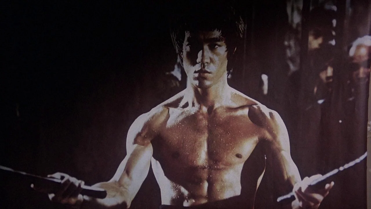 Saturday Night Fever (1977) movie still: medium — An dimly lit interior space with indistinct dark surroundings. A muscular man holds nunchaku in…