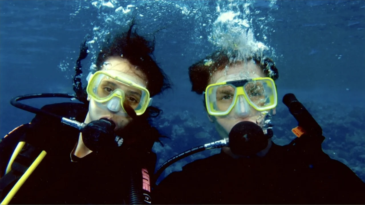 The (Dead Mothers) Club (2014) movie still: medium — Two scuba divers hover submerged beneath the surface of clear blue water. They gaze directly…