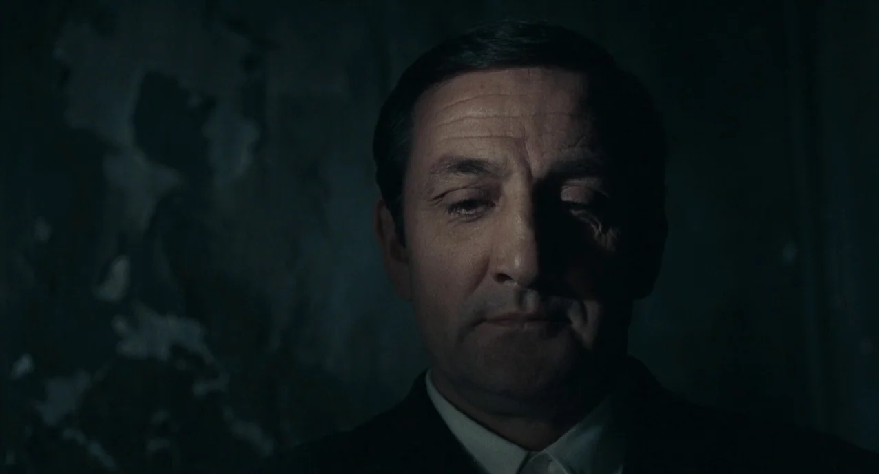 The Army of Shadows (1969) movie still: closeup — A man stands in a dimly lit, dark-walled space with textured, peeling paint. He looks toward the…